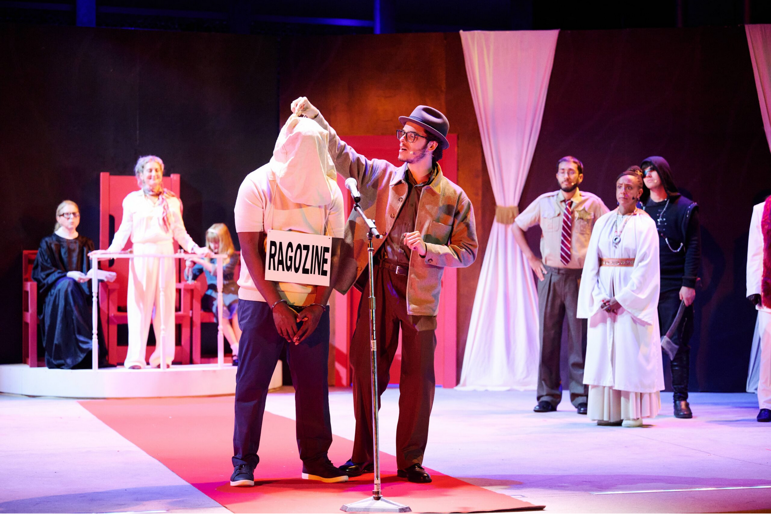 Ragozine stands in handcuffs and a man begins to remove the hood from his head. They stand in front of a judging panel.